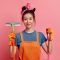 Cheerful housewife with cleaning tools on pink background.